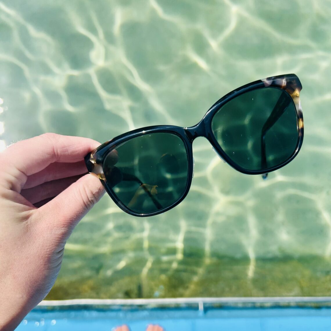 Sonnenbrille von einer Hand gehalten, Schwimmbecken im Hintergrund